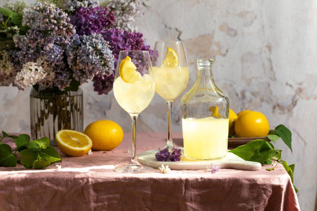 Zwei Gläser gefüllt mit sprudelnder Zitronenlimonade, garniert mit Zitronenscheiben, neben einer Karaffe auf einem Tisch mit einem Blumenstrauß im Hintergrund, betont durch Teisseire Sirup für erfrischenden Geschmack.