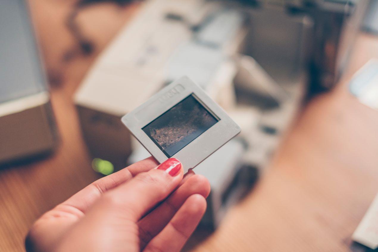 Eine Hand hält ein Dia, während im Hintergrund ein Scanner steht – ideal für den DIY-Prozess zum Dias digitalisieren.